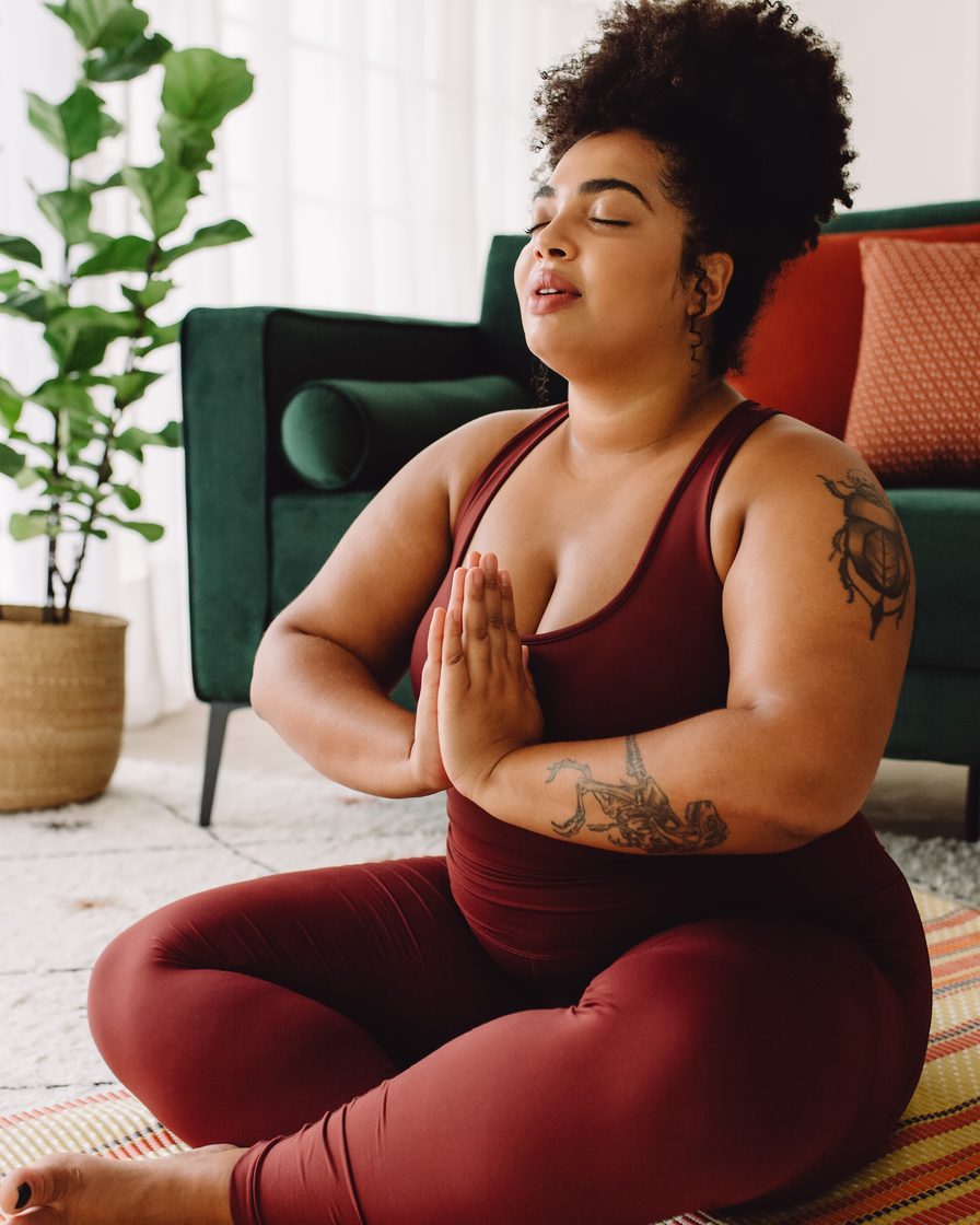 A woman sitting cross-legged on a yoga mat at home, hands at heart center and eyes closed in quiet meditation